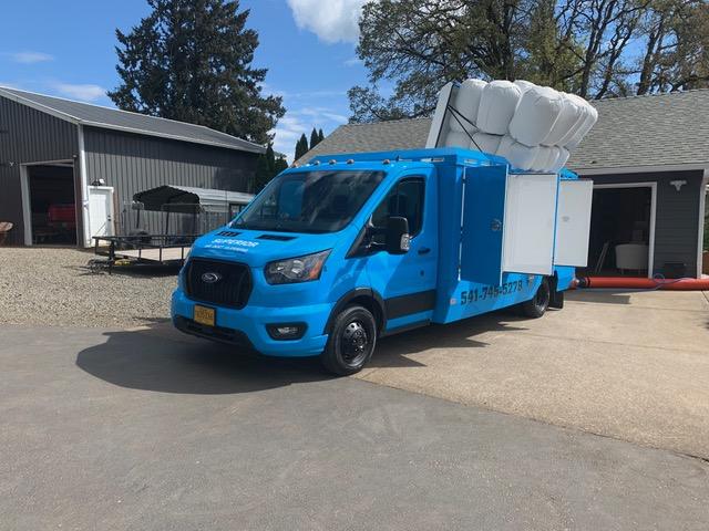 Blue air duct cleaning box truck with its vacuum hoses raised, parked outside a workshop